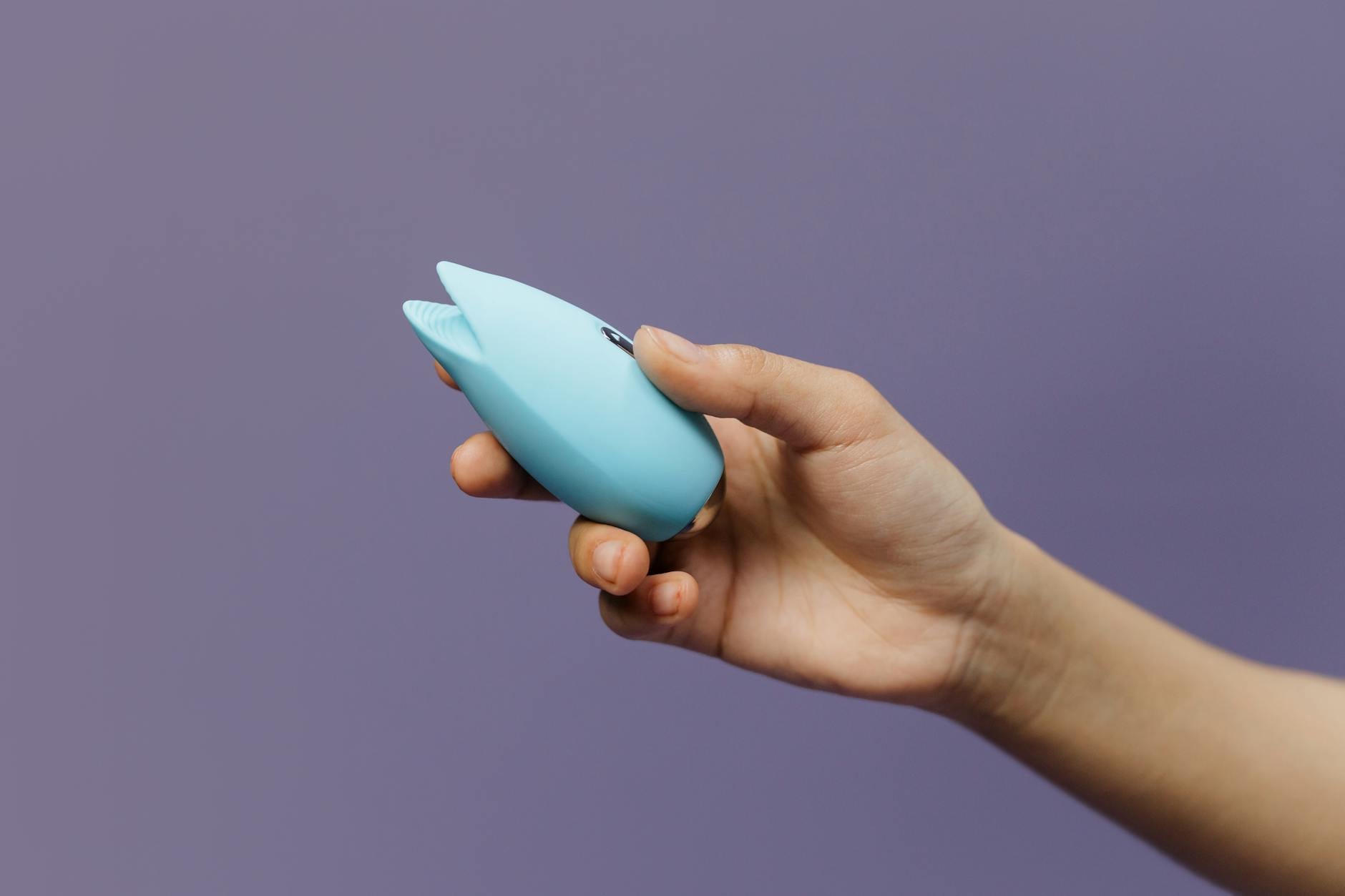 A hand holding a blue silicone clitoral vibrator against a purple background, representing self-love and intimate pleasure
